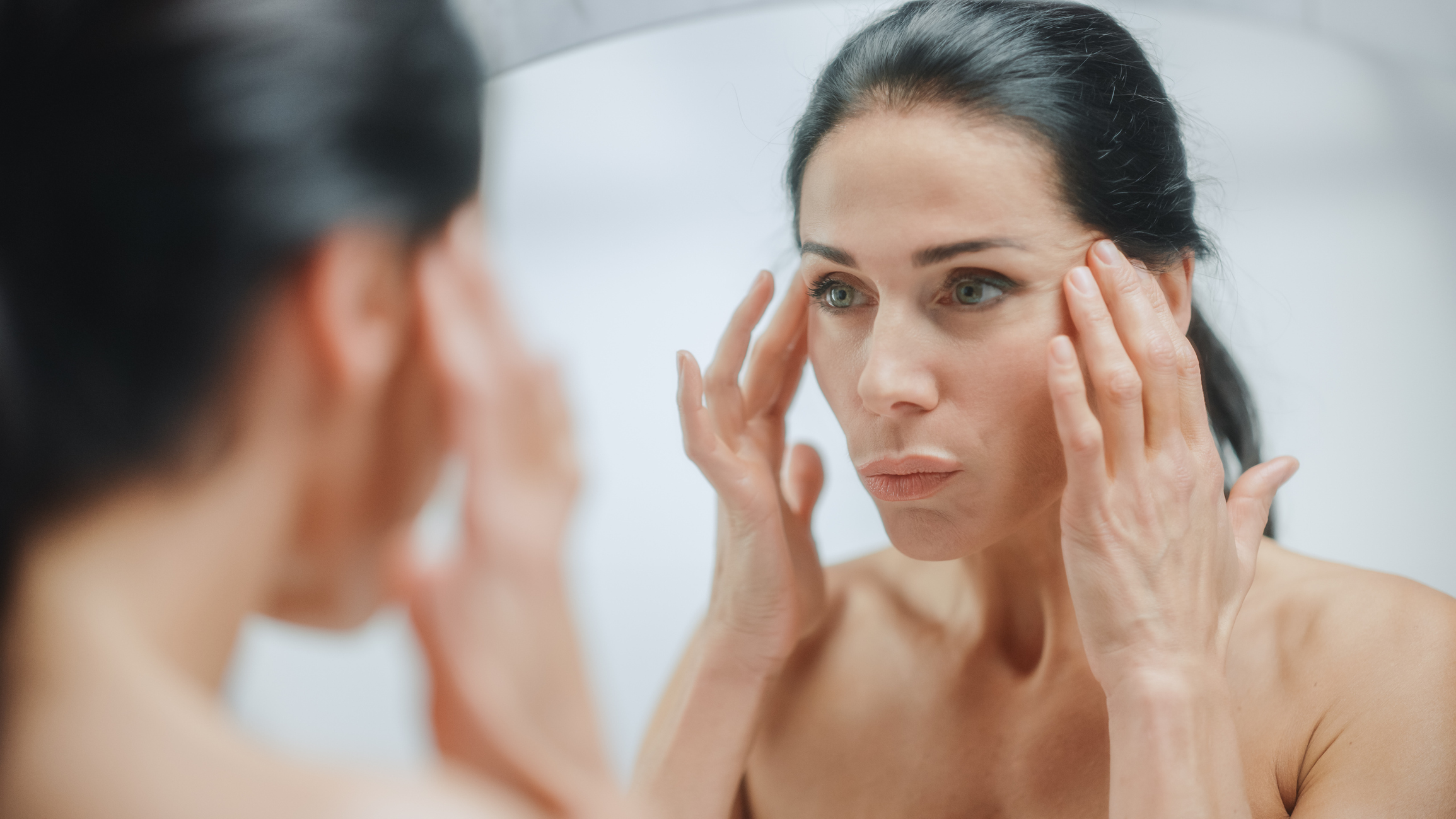 Caucasian Woman Looking in Mirror, Touching Beautiful Face, Soft Skin. Elderly Lady with Lush Black Hair. Female Beauty, Dignity, Grace. Sensual Wellness and Organic Skincare Products. Portrait Shot Eyelid Surgery in McAllen, TX